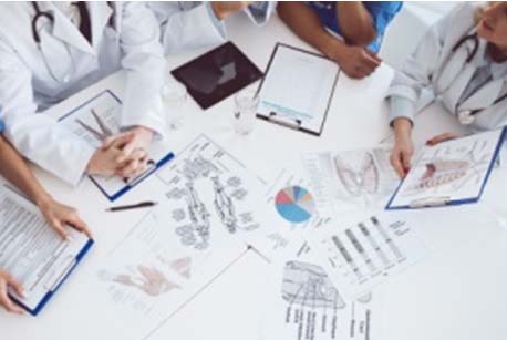 Instantània on es veuen doctors i doctores en bata blanca amb actitud de comentar els informes que hi ha sobre d'una taula.